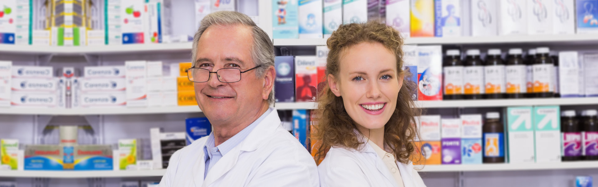 Pharmacists smiling while being side by side with a lot of medicine in the background